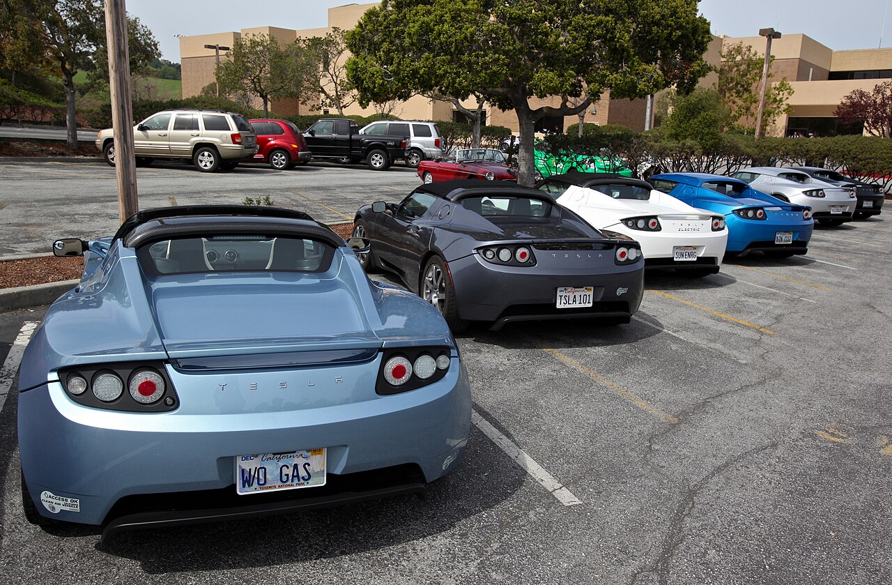 Several first-generation Tesla Roadsters in light blue, gray, white and blue, parked in a row in a parking lot, seen from behind