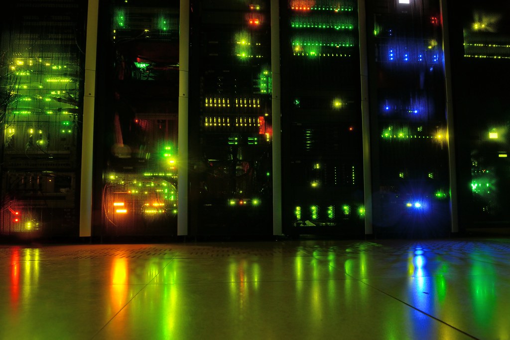 A row of tall black server racks glowing with green, blue, yellow and red indicator lights, reflected on a shiny tiled floor.