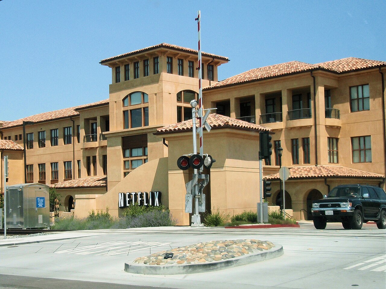 A three-story tan building with a red tile roof and a Netflix sign, with a railroad crossing signal in front